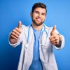 Young blond doctor man with beard and blue eyes wearing white coat and stethoscope approving doing positive gesture with hand, thumbs up smiling and happy for success. Winner gesture.